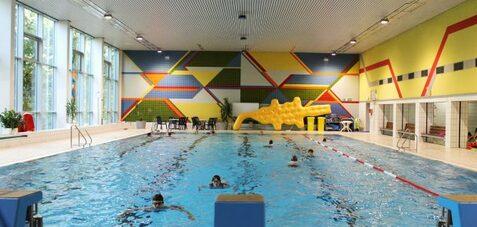 Schwimmbecken in einem Hallenbad mit bunten Wänden und mehreren Personen im Wasser. Rutschbahn an der Seite.
