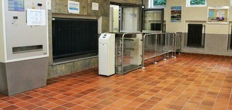 Menü mit Ticketautomat und Drehkreuz in einem hellen Innenraum mit Fliesenboden und Fenster.