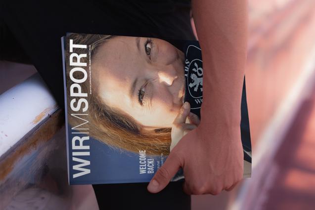 Nahaufnahme einer Person, die ein Magazin mit dem Titel „WIR IM SPORT“ in der Hand hält, im Hintergrund eine Sportfläche.